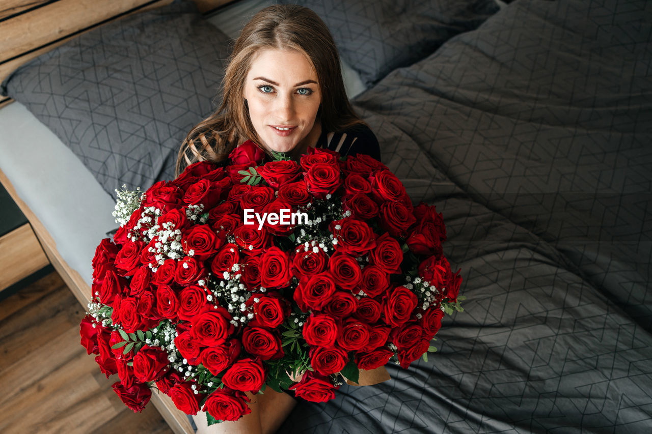 A beautiful girl is sitting on a bed in the bedroom with a huge bouquet of scarlet roses. 