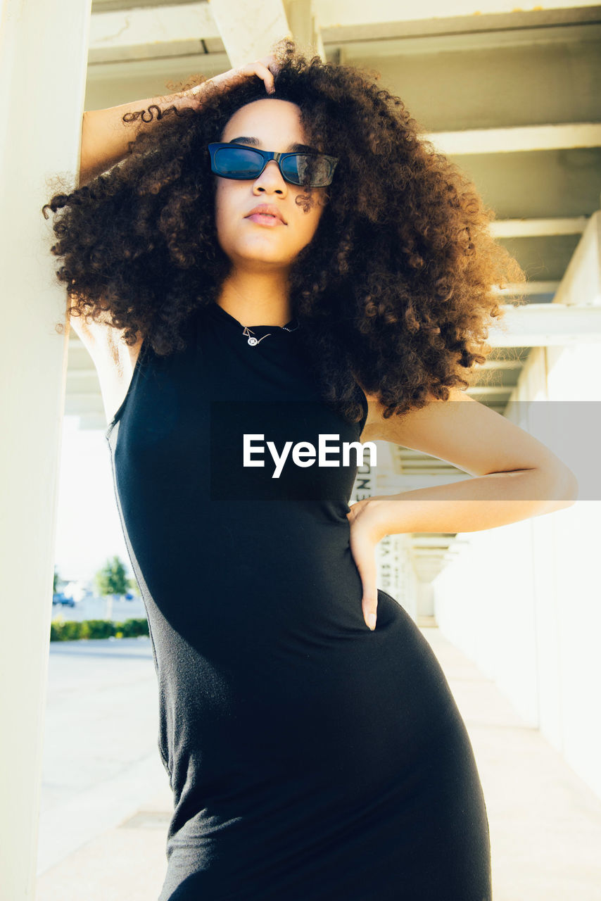 Cool young ethnic woman with afro curls wearing black dress and sunglasses standing near stone column with hand on waist under roof near building in sunny day in street