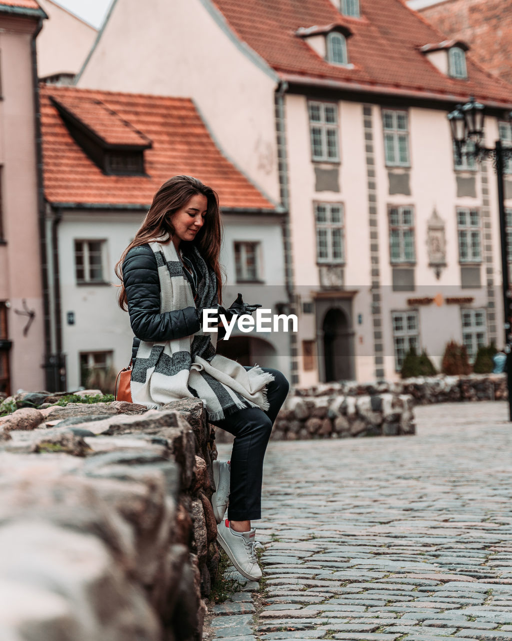 Young woman sitting on stone wall against buildings