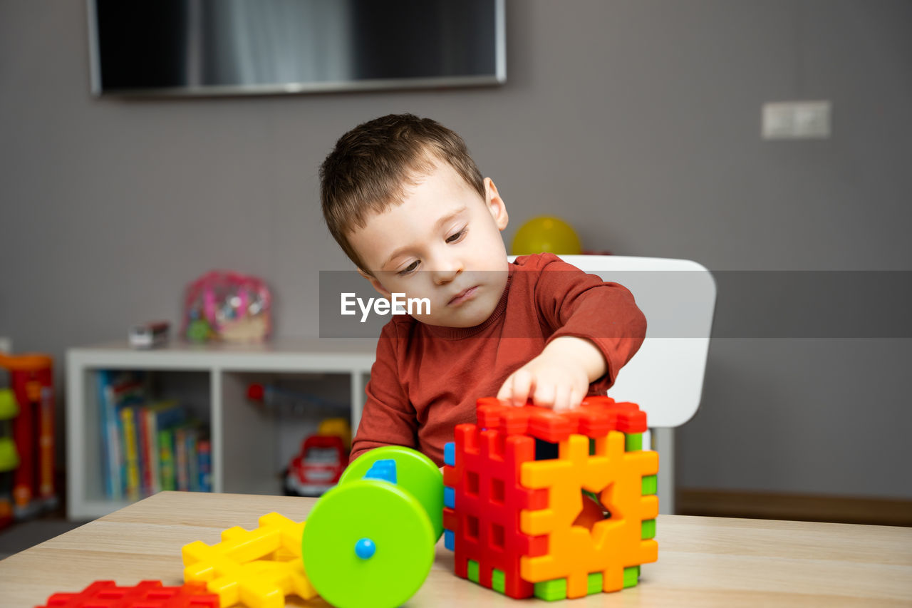 A cute attentive little toddler boy of two years old sits at a children's table and plays