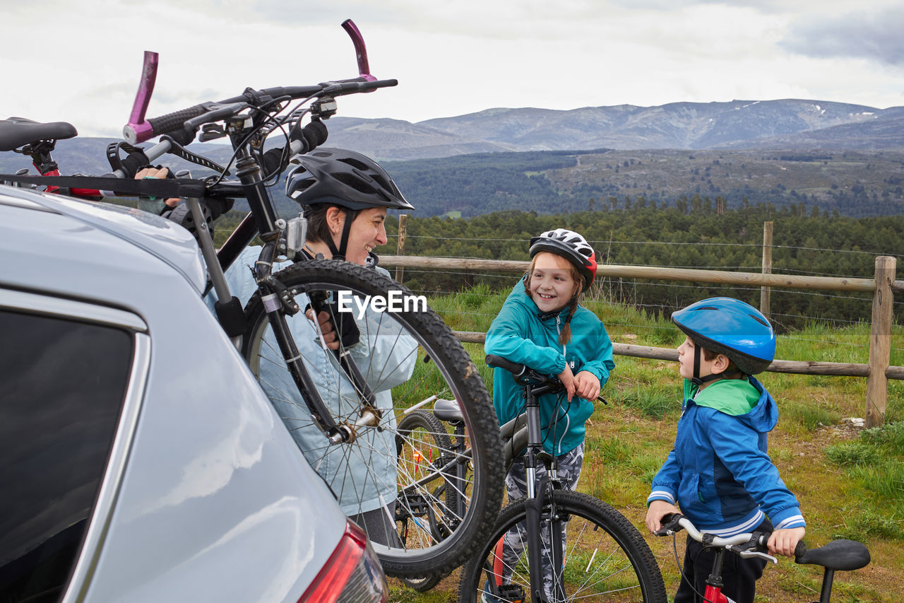 Side view of positive mother with kids in helmets taking bicycle from trunk rack of modern car parked in countryside