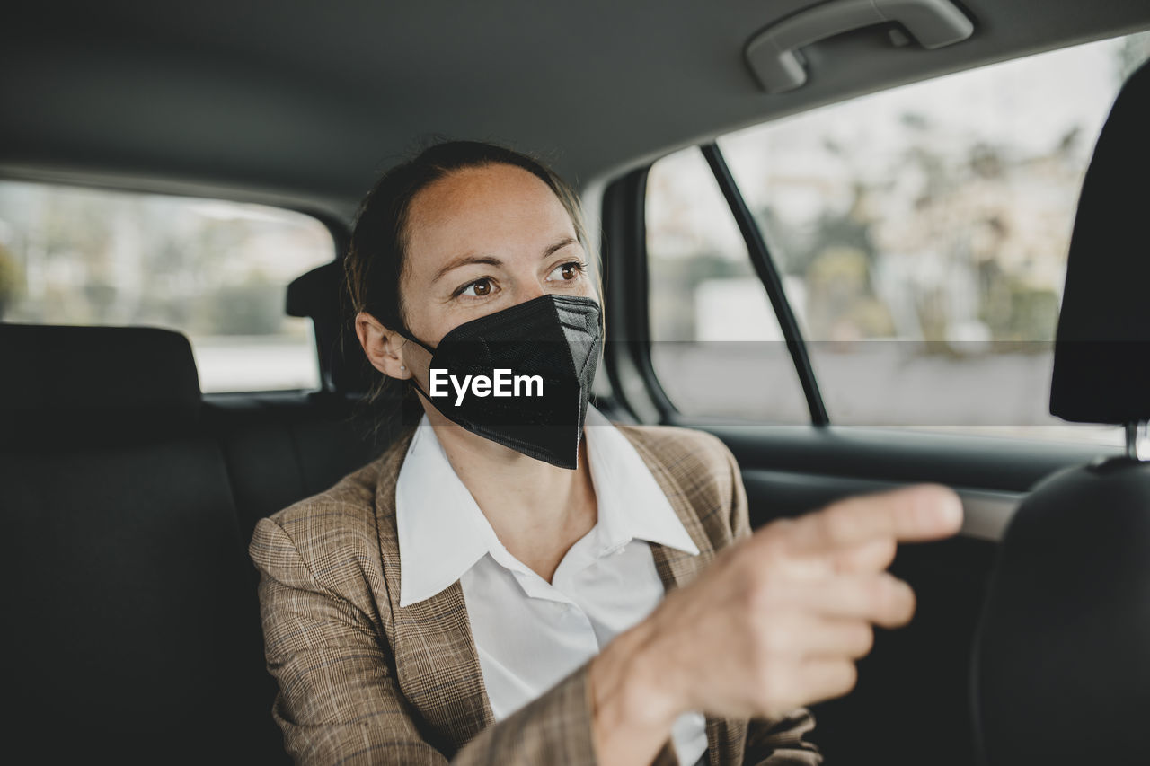 Female professional in face mask gesturing while traveling in taxi during covid-19