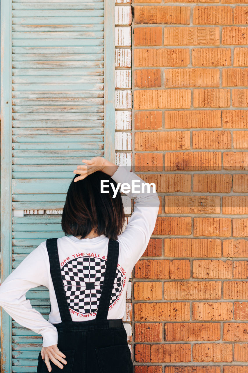 REAR VIEW OF SENIOR WOMAN STANDING AGAINST BRICK WALL