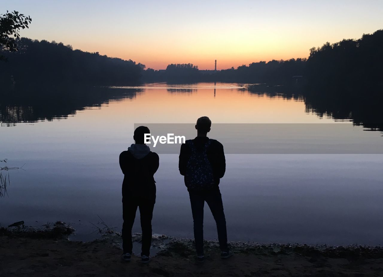 Rear view of silhouette men standing by lake against sky during sunset