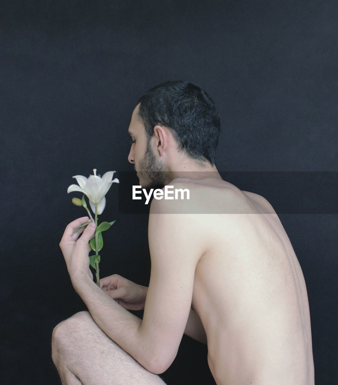Side view of shirtless young man holding flower while crouching against wall