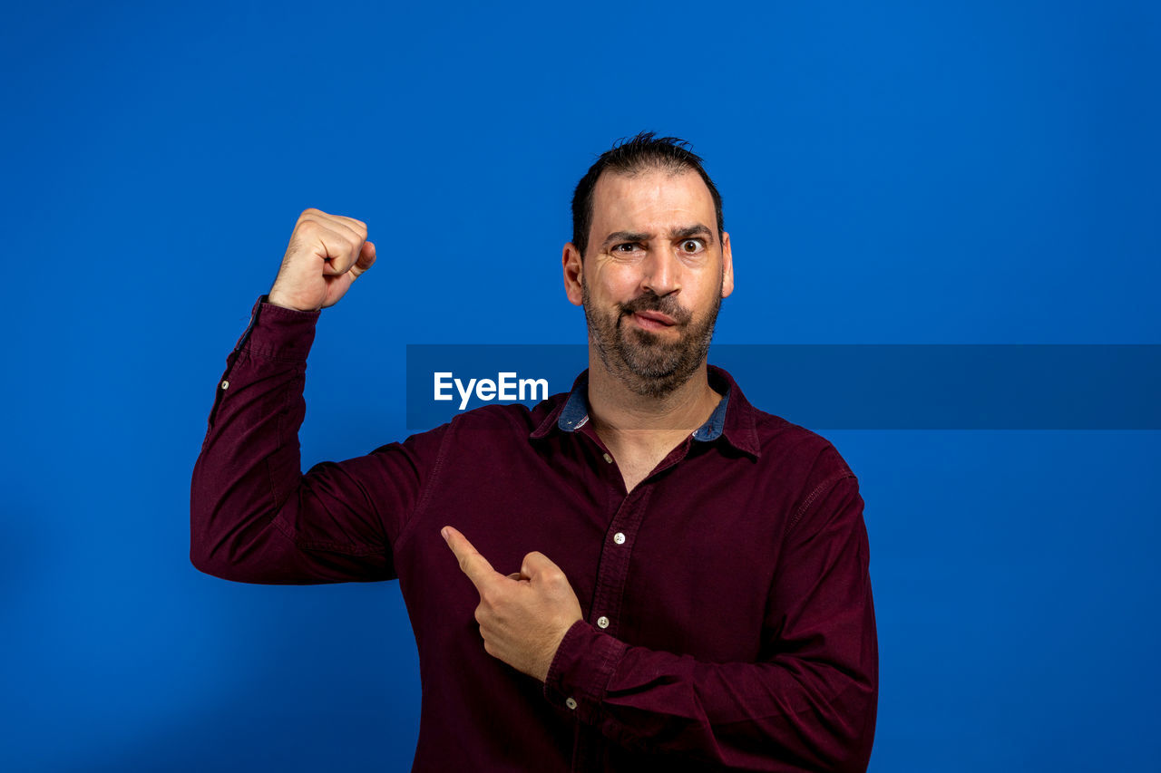 blue, one person, adult, men, portrait, blue background, studio shot, colored background, looking at camera, waist up, beard, person, smiling, facial hair, emotion, happiness, front view, sign language, standing, indoors, casual clothing, copy space, finger, gesturing, success, positive emotion, cheerful, hand, human face, arm, business