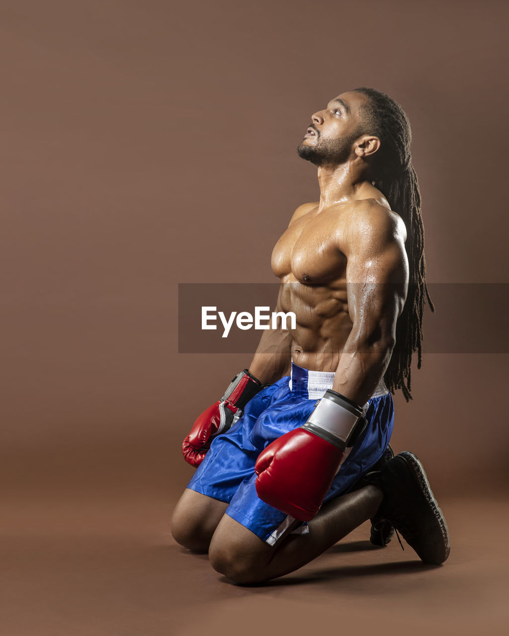 Full length of shirtless boxer kneeling on brown background