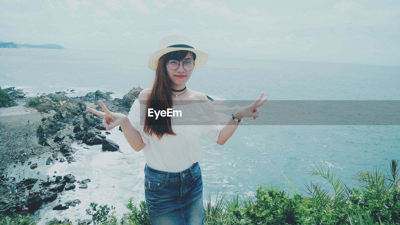 Portrait of woman gesturing peace sign while standing against sea