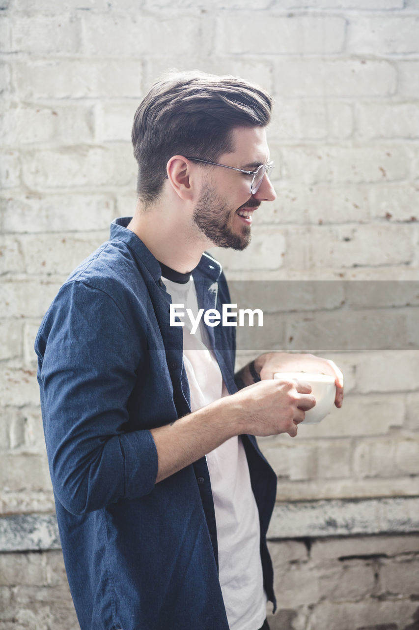 Side view of bearded smiling computer programmer holding coffee cup while standing in office