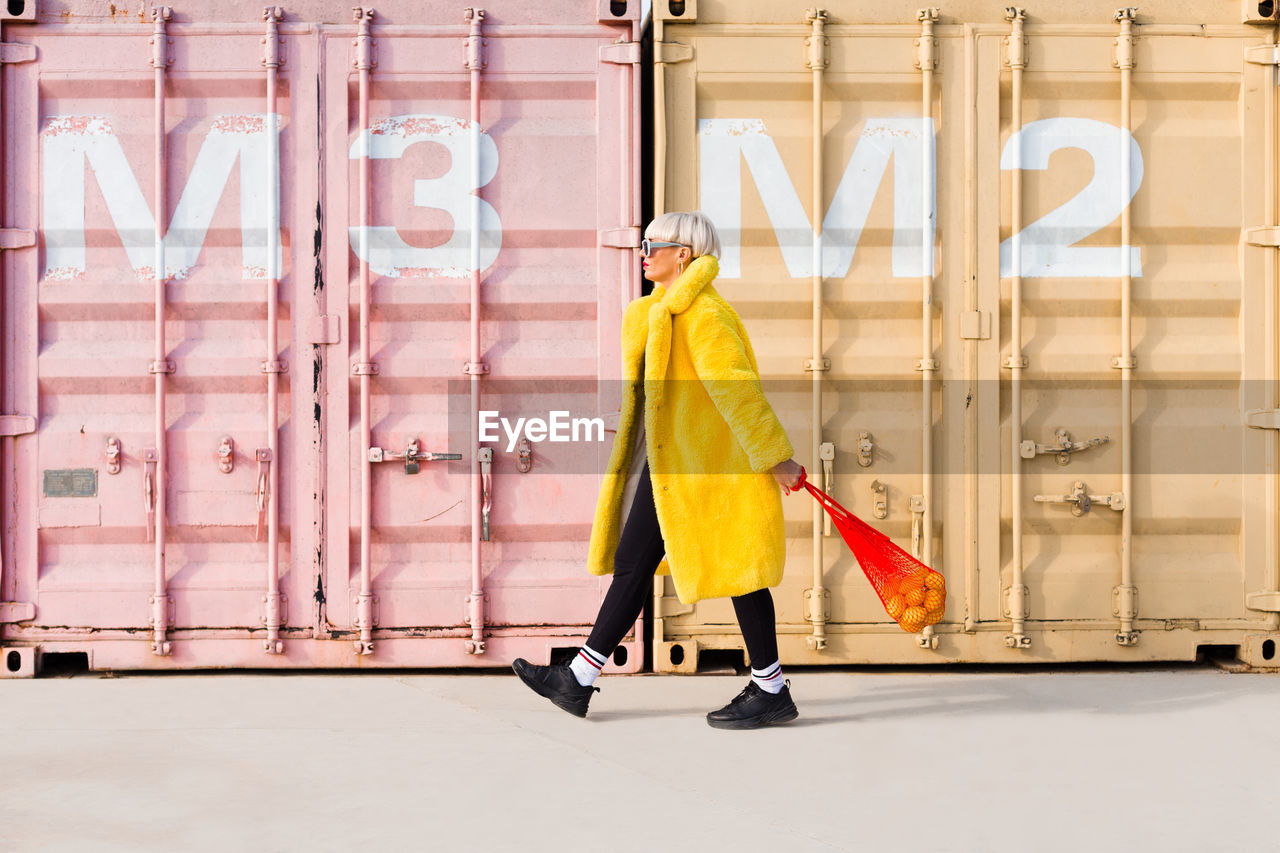 Side view of trendy female hipster in vibrant outfit walking along street with sack full of oranges and looking away