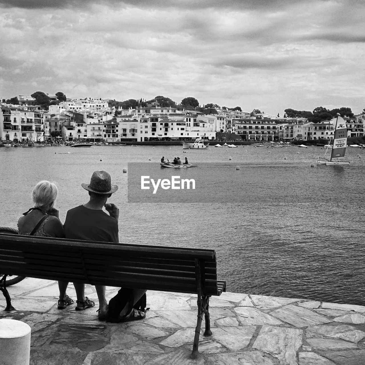 Man and woman sitting on bench looking at boats in river by city