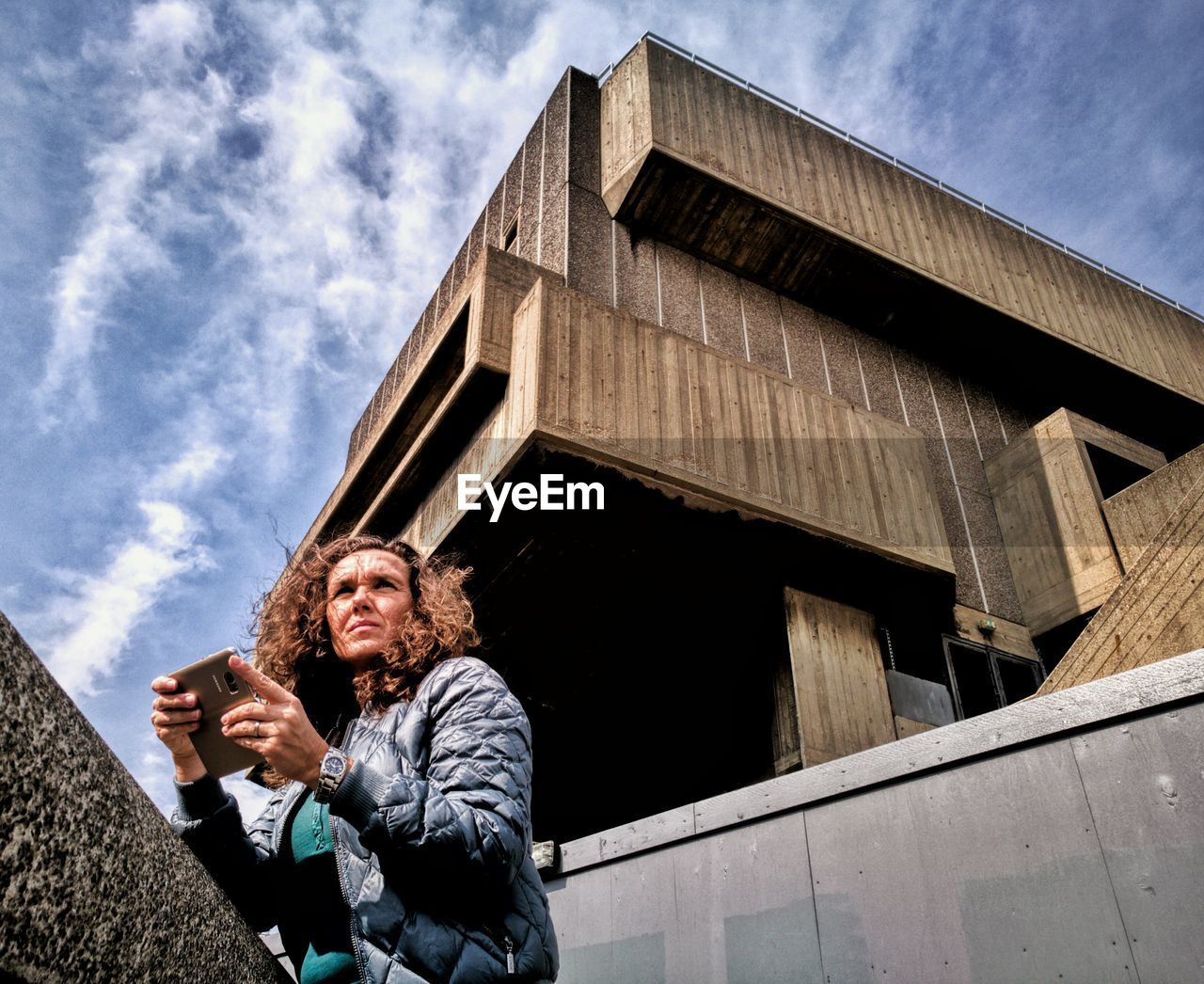 Low angle view of woman using mobile phone while standing by retaining wall against cloudy sky