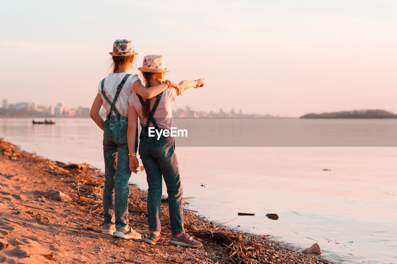 Two girls sisters stand on the banks of the river at sunset and look into the distance