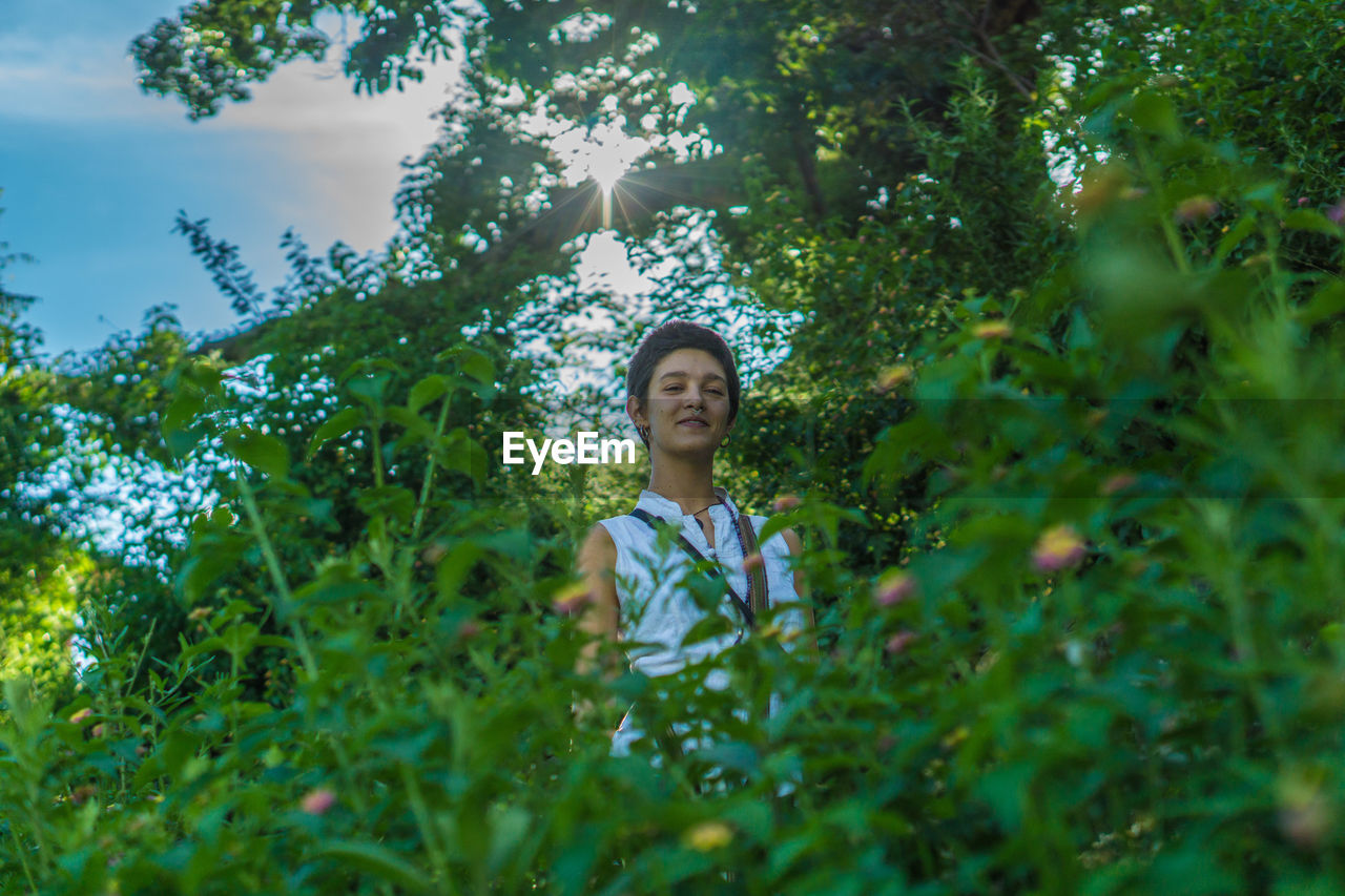 Portrait of young woman standing by plants against trees