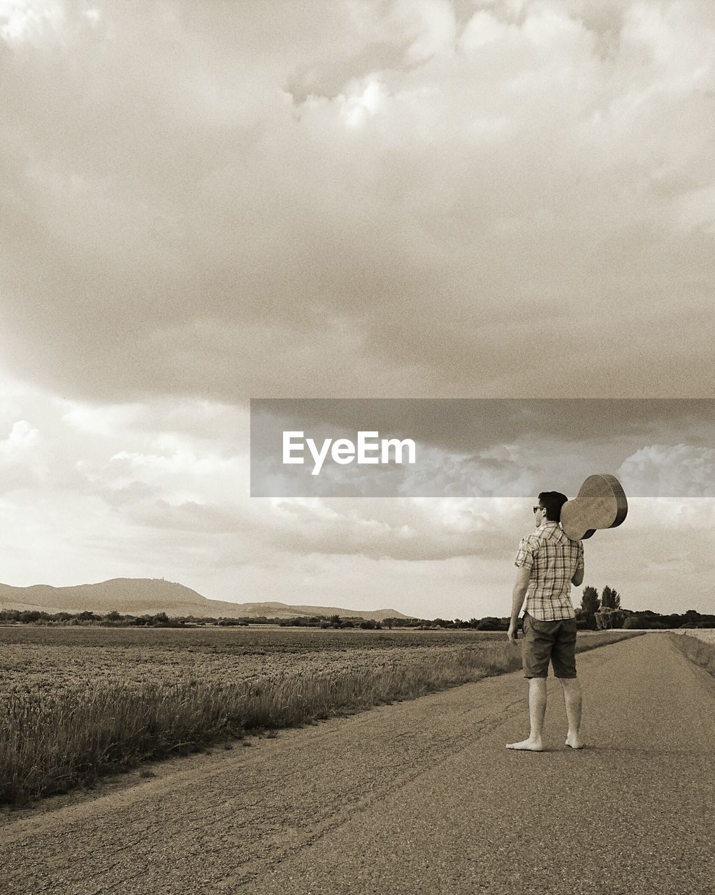 Rear view full length of man with guitar standing on road by field against sky