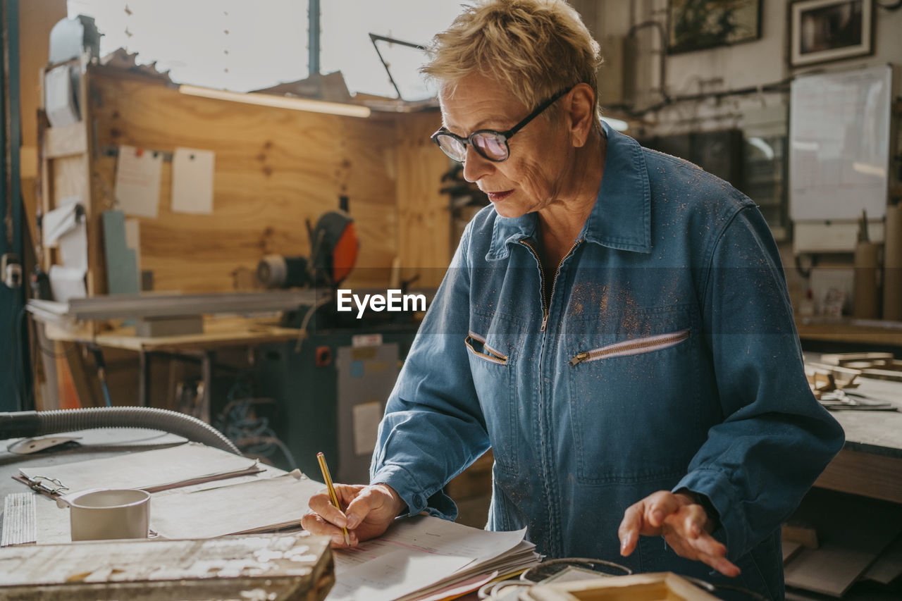 Senior female carpenter wearing eyeglasses preparing document at workshop