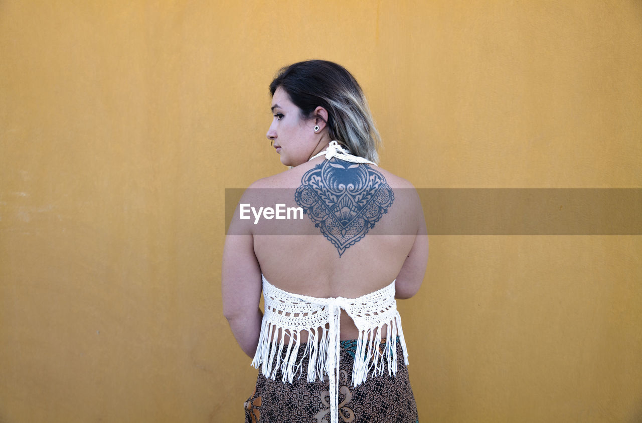 Rear view of young girl showing her tattoo in back against yellow wall