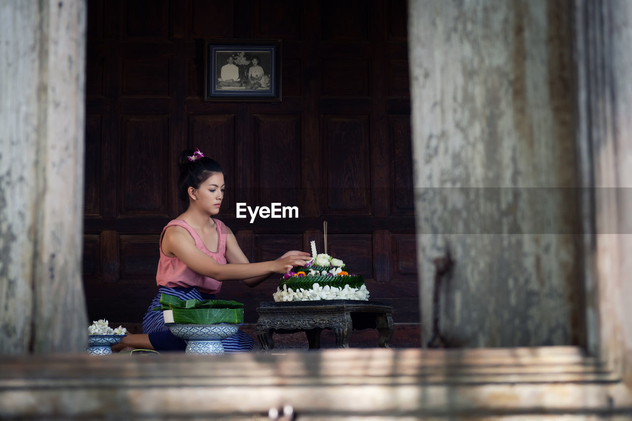 Young woman arranging flowers on table at home seen through window