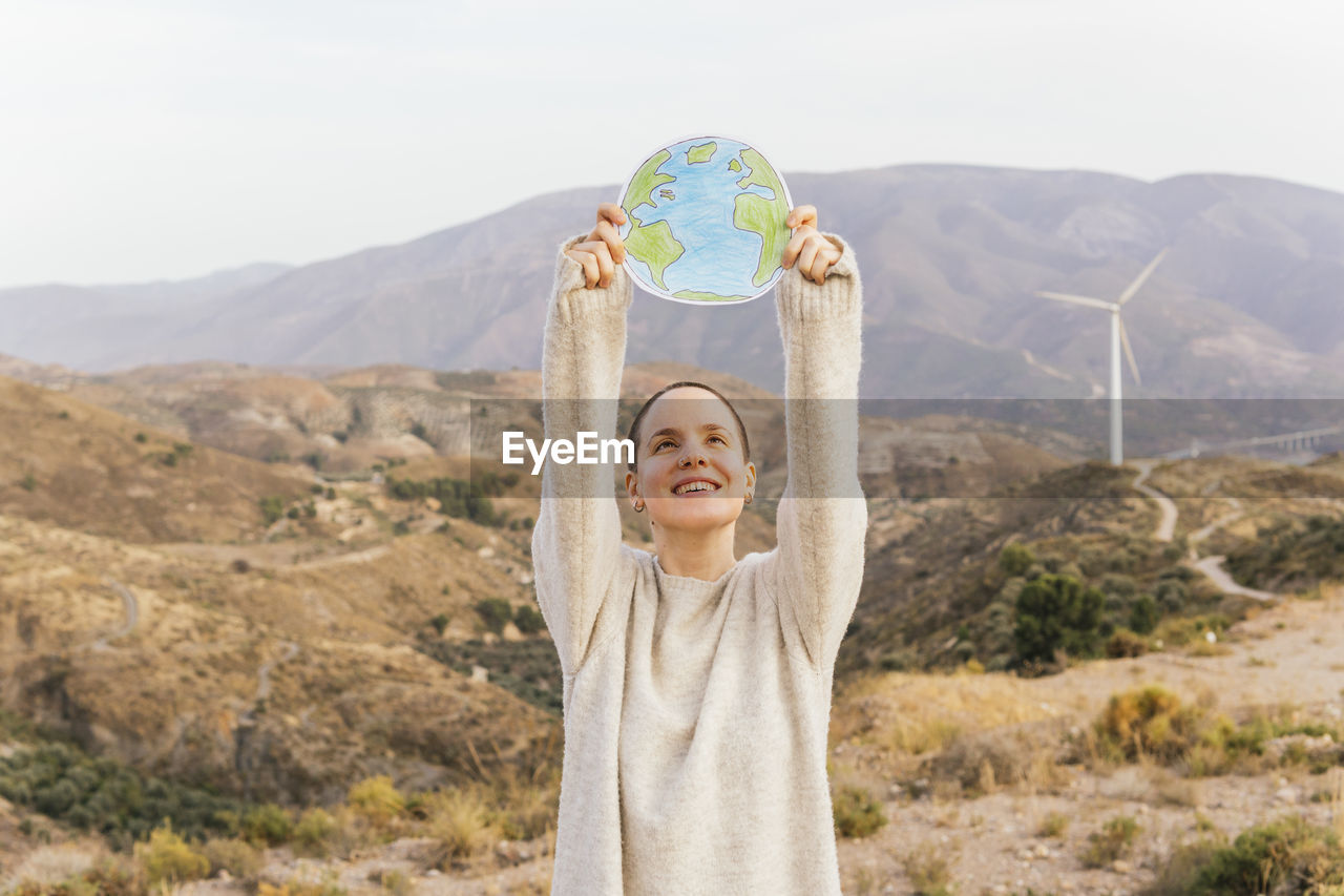 Smiling shaved head woman with hand raised holding planet earth on mountain