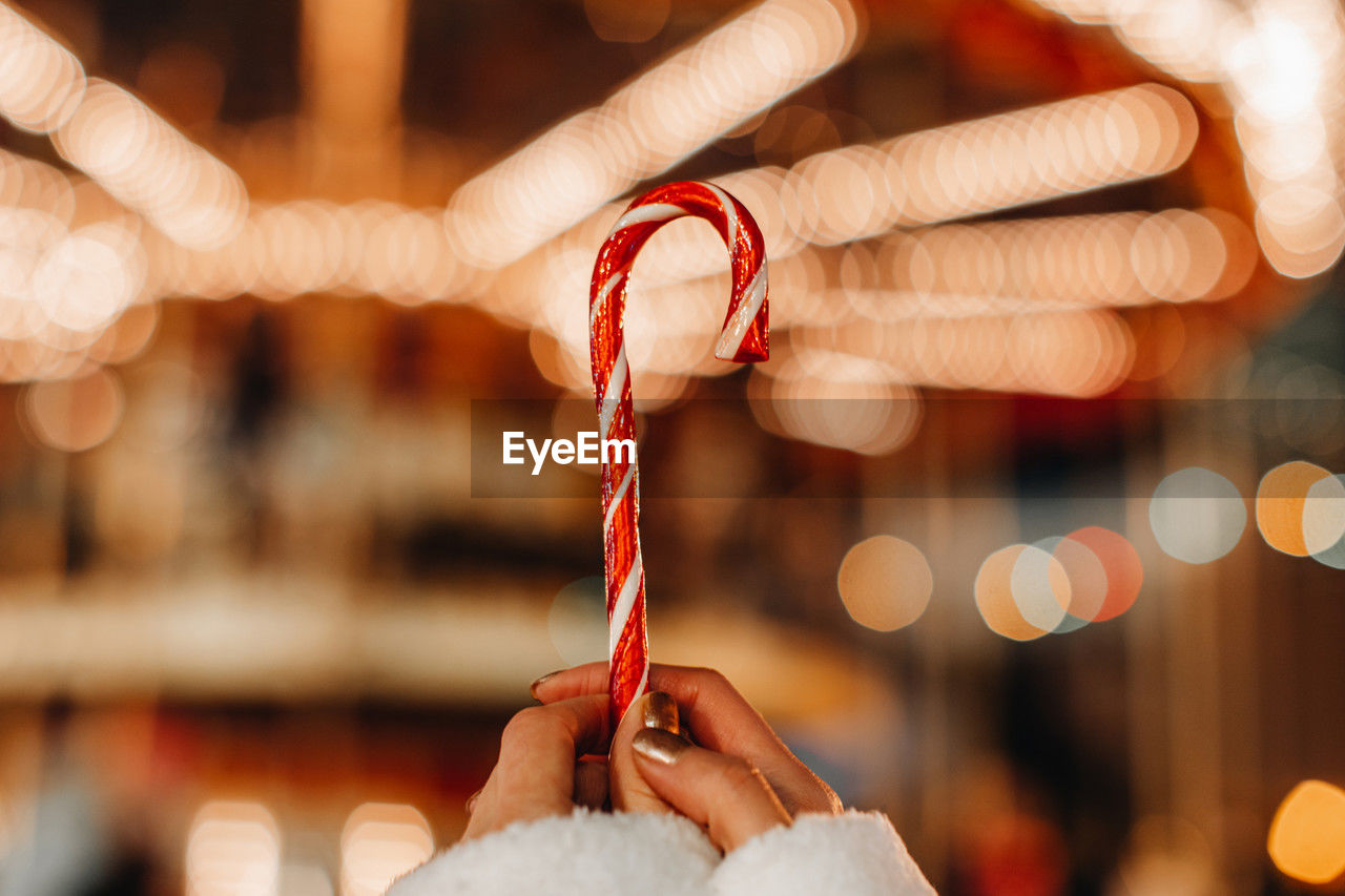 Female hands holding red white cane sweet lollipop against the background of golden garland lights