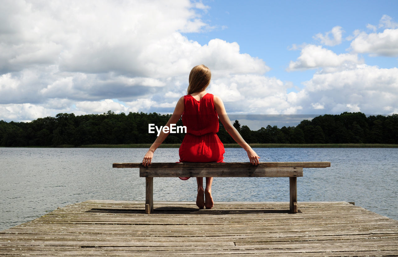 Rear view of woman sitting on bench by lake against cloudy sky