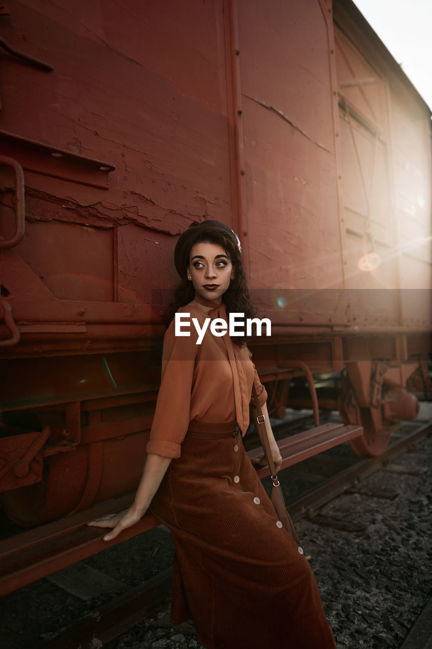 Young female with dark curly hair in beret wearing terracotta clothes in vintage style in back lit sitting on step of car train painted in terracotta color and holding handrail while looking away flirty