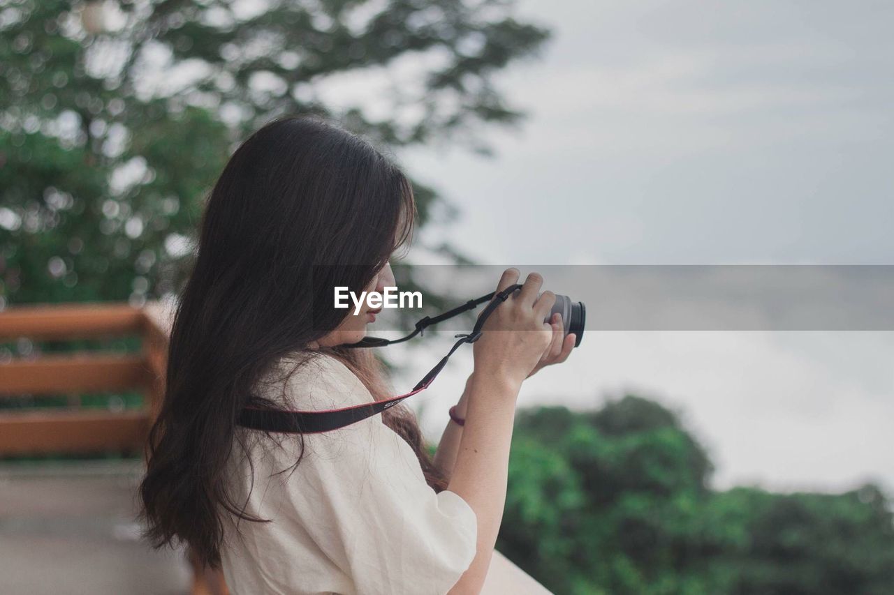 Side view of woman holding camera against sky