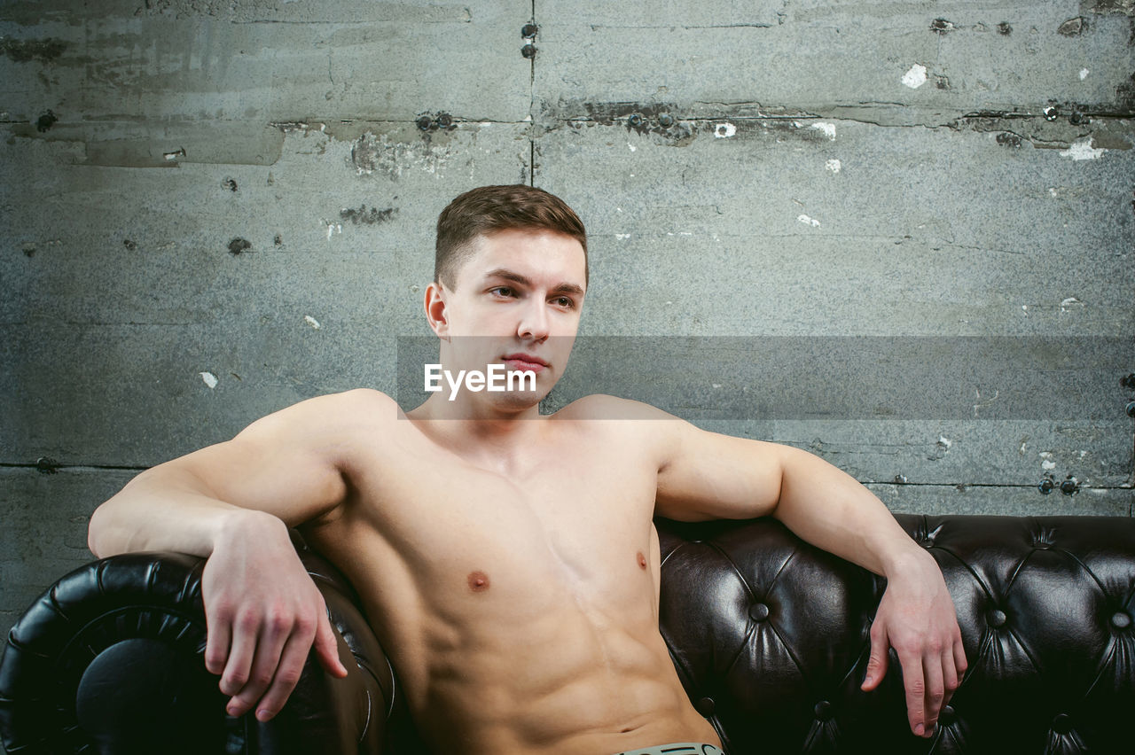 Shirtless muscular man sitting on armchair by wall