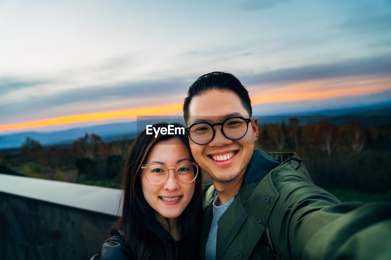 Portrait of smiling couple taking selfie against sky during sunset