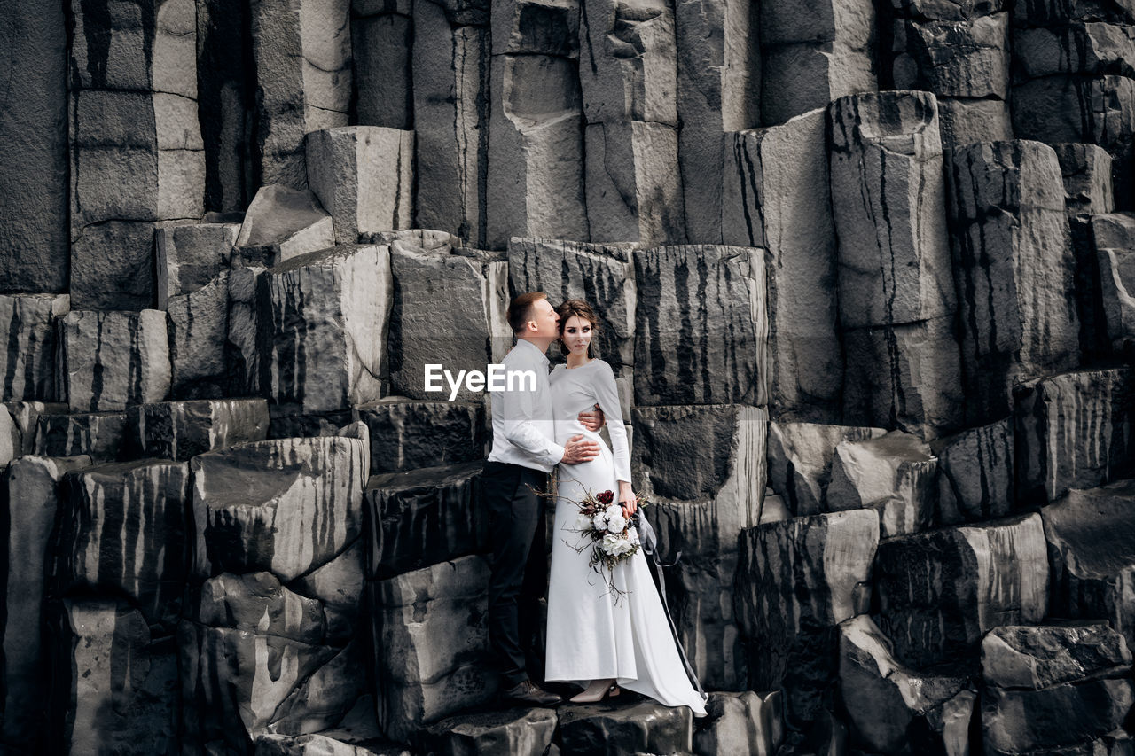 FULL LENGTH OF COUPLE STANDING IN FRONT OF TRADITIONAL CLOTHING