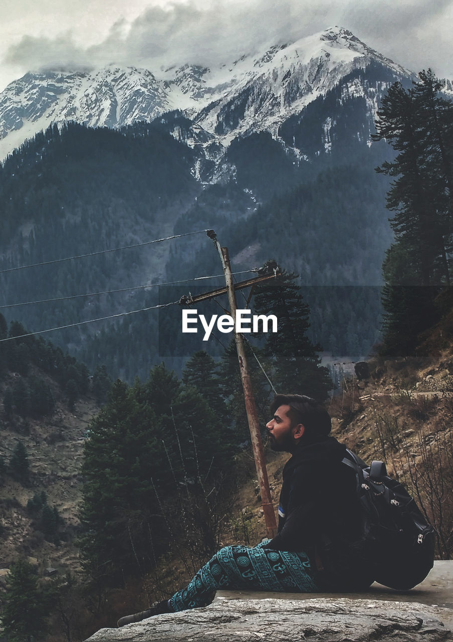 Young man sitting on rock against snowcapped mountains