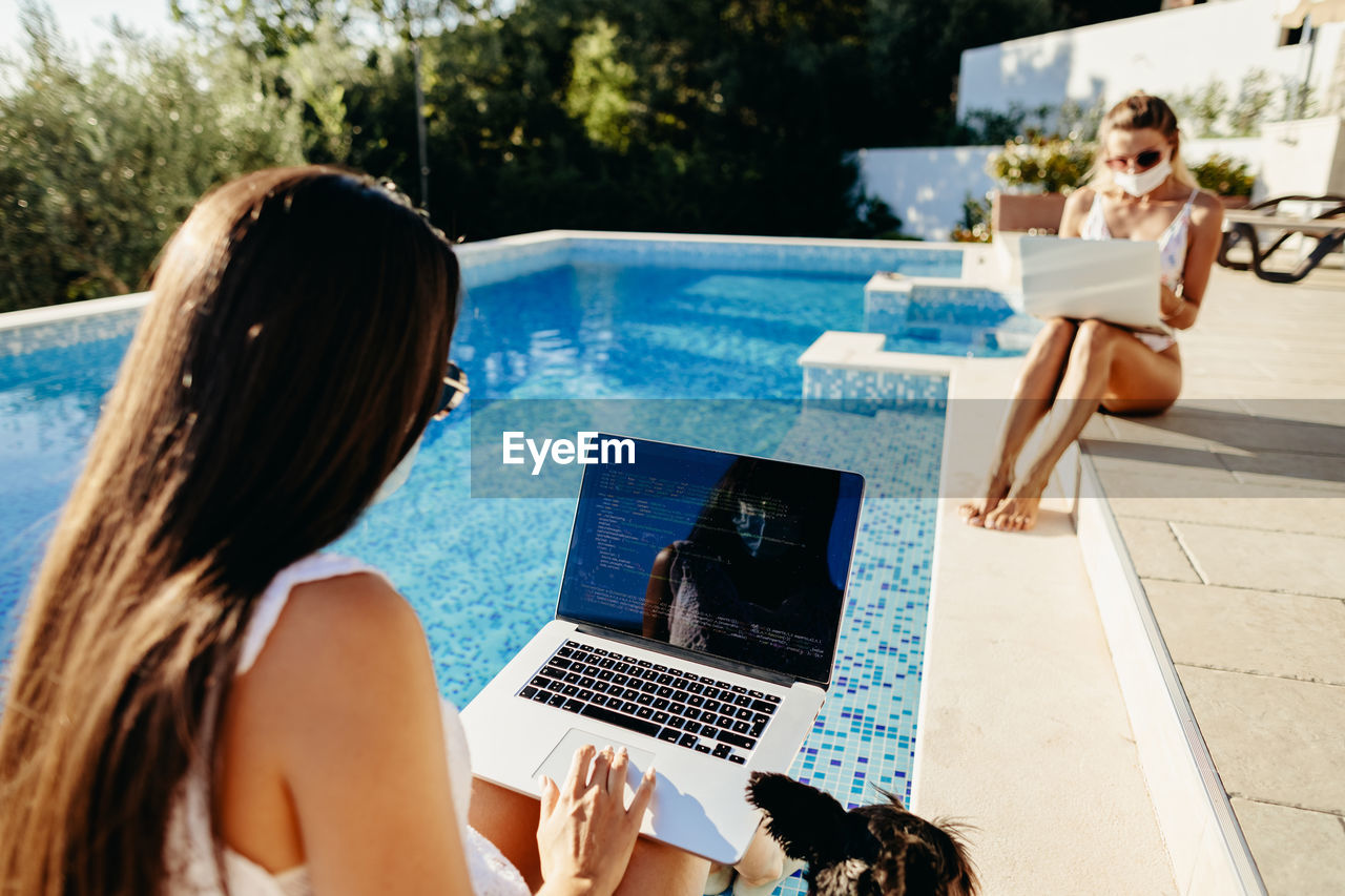 Rear view of woman using laptop sitting by swimming pool