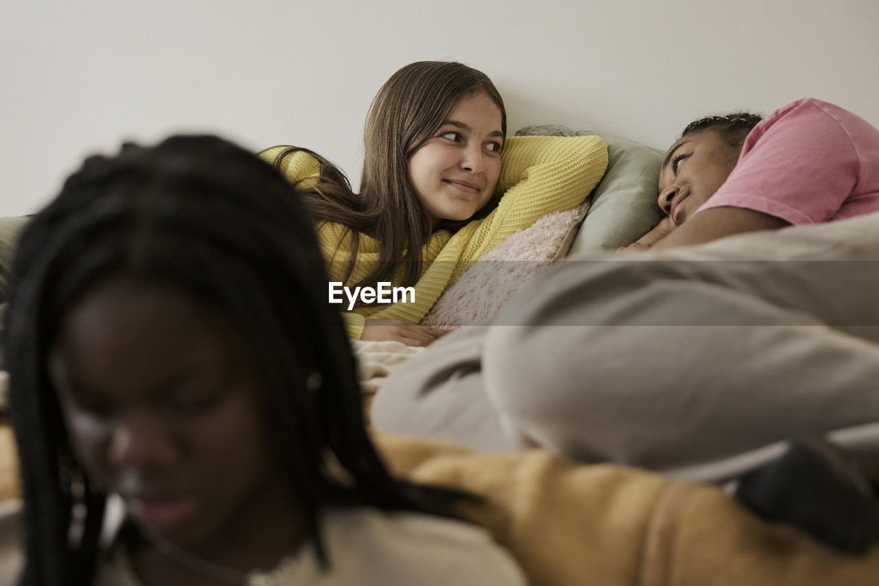 Teenage girls talking while lying in bed by female friend sitting in front at home