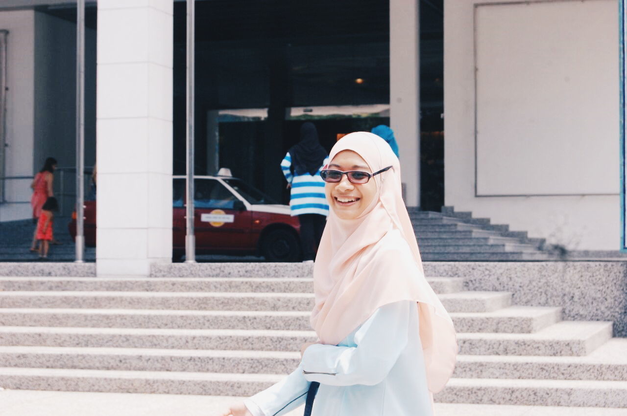 PORTRAIT OF SMILING YOUNG WOMAN STANDING AGAINST WALL