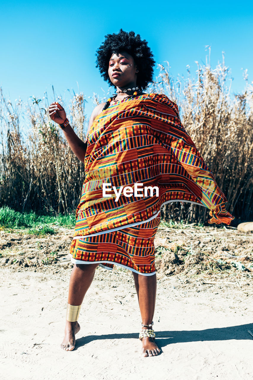 Serious african model with afro hairstyle wearing colorful handwoven kente and looking away while standing on field on bright summer day