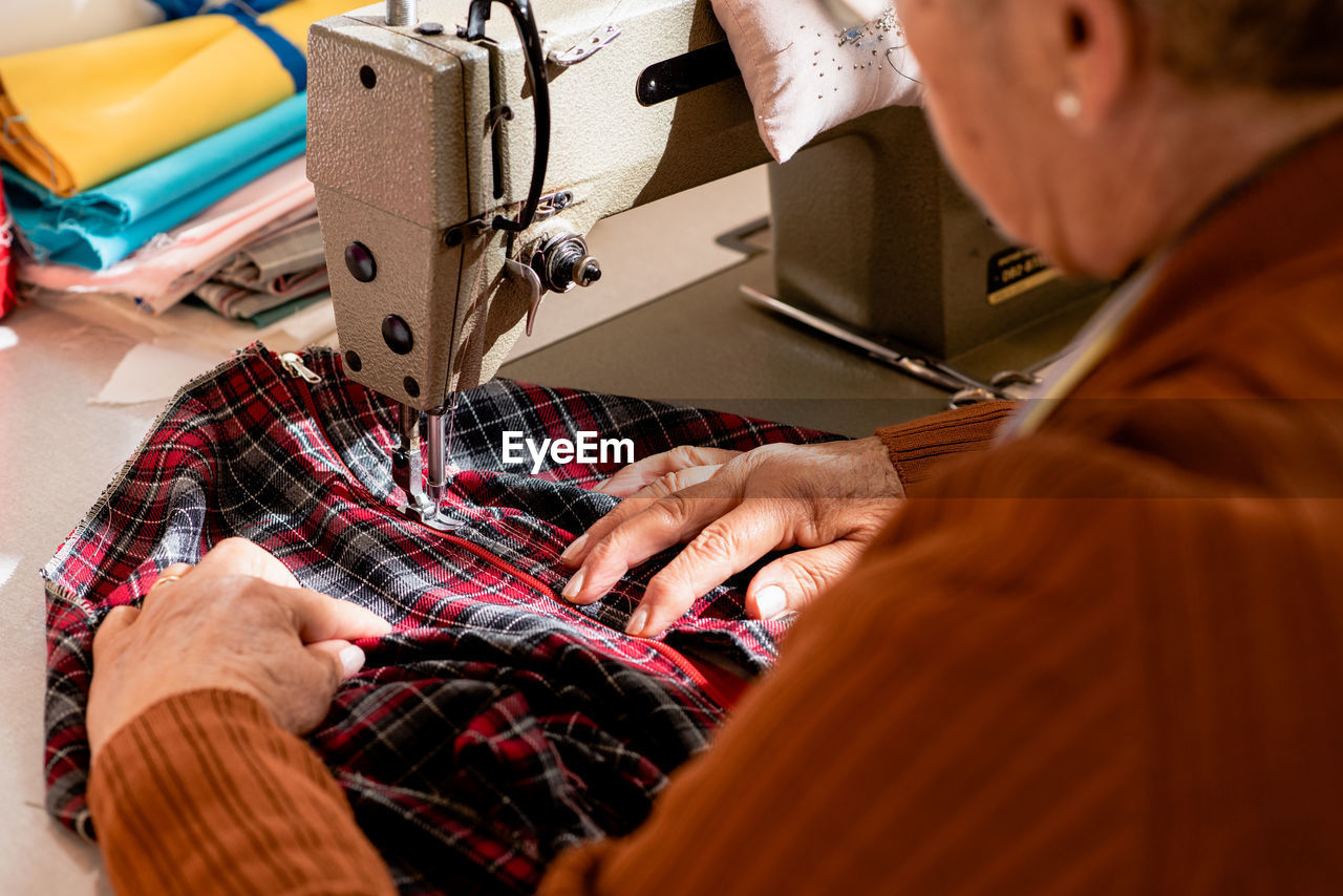Cropped unrecognizable senior seamstress sitting at sewing machine and stitching piece of cloth while working in workshop