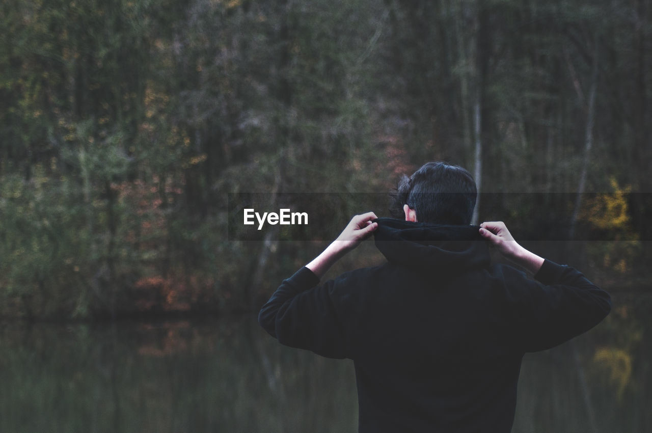 Rear view of man wearing hooded shirt by lake in forest