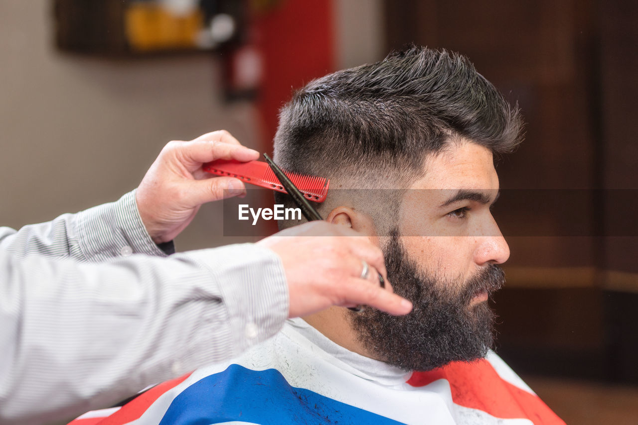 Cropped hands of barber cutting male customer hair with scissors in salon