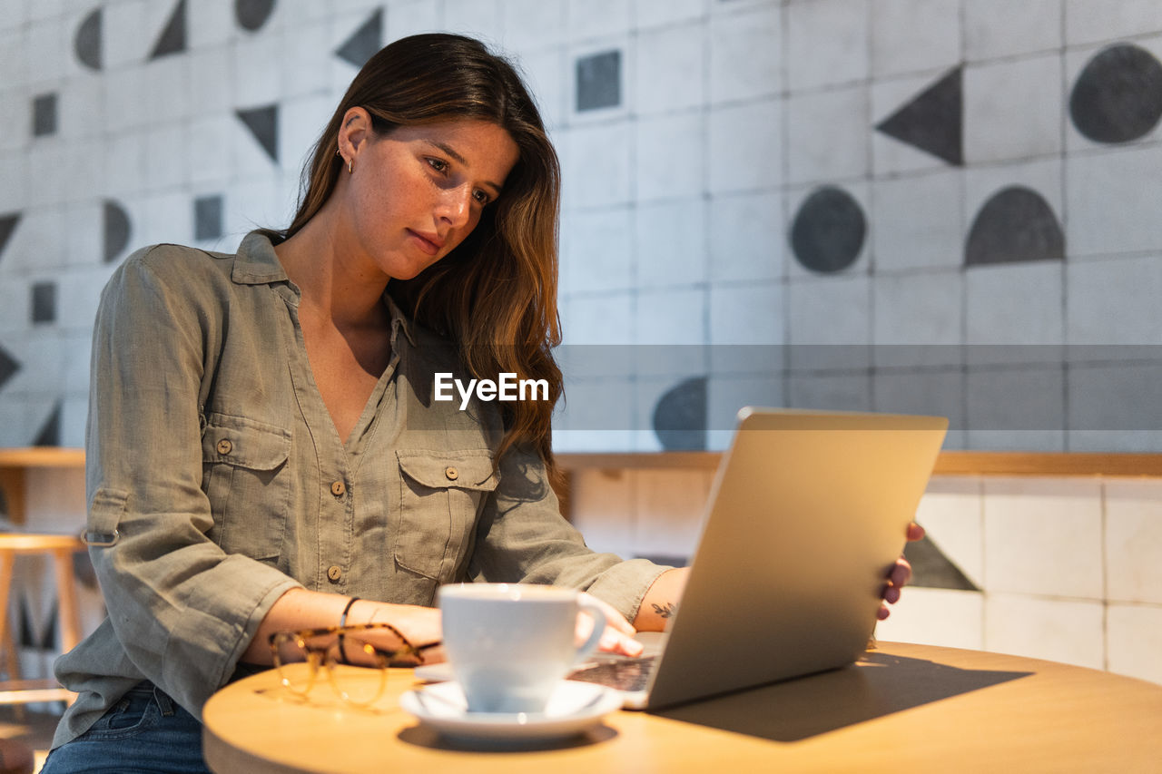 Young focused female browsing internet on portable computer at table with hot drink in cafeteria