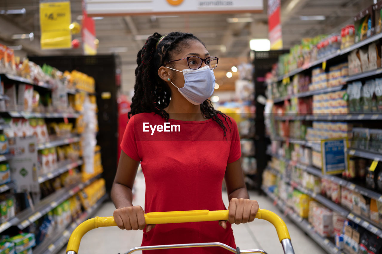 Afro latina young woman wearing a face mask pushes shopping cart through supermarket aisle