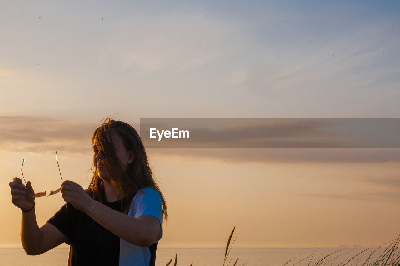 Smiling woman holding sunglasses standing against sky