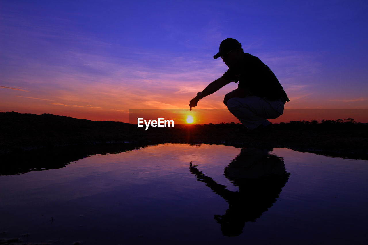 Optical illusion of man touching sun while crouching by water during sunset