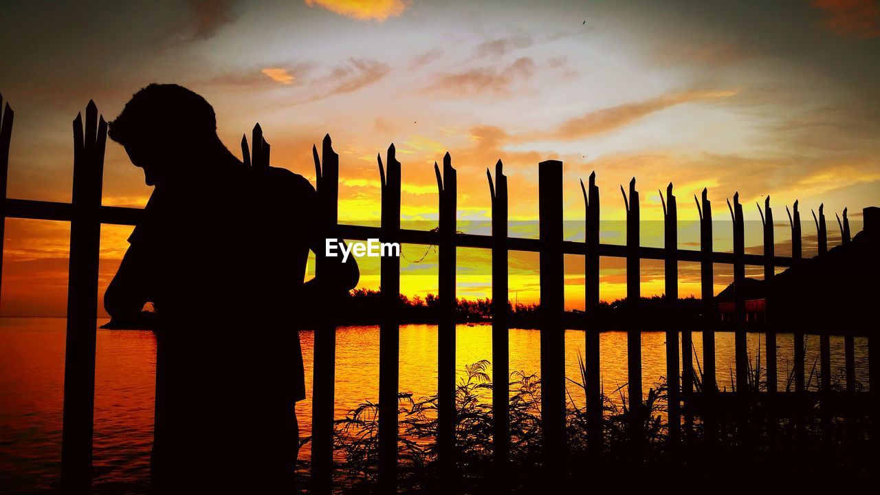 Silhouette man standing by railing in front of sea against sky at dusk