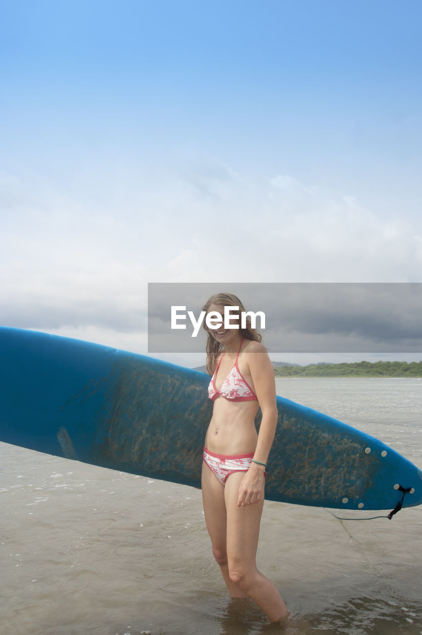 Happy woman in bikini with surfboard standing at sea against sky