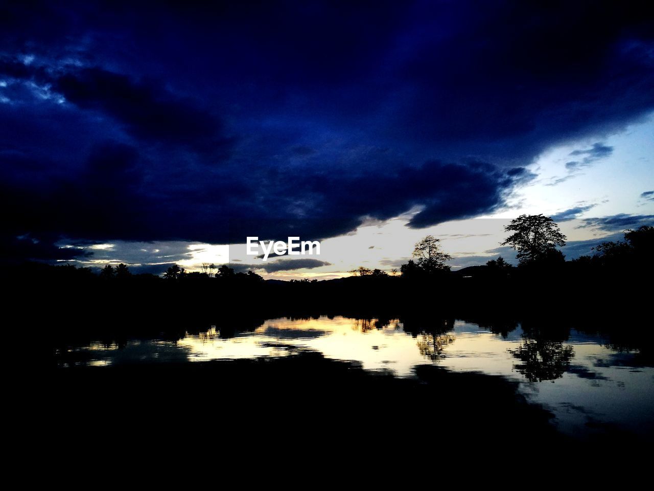 REFLECTION OF SILHOUETTE TREES IN LAKE AGAINST SKY AT SUNSET