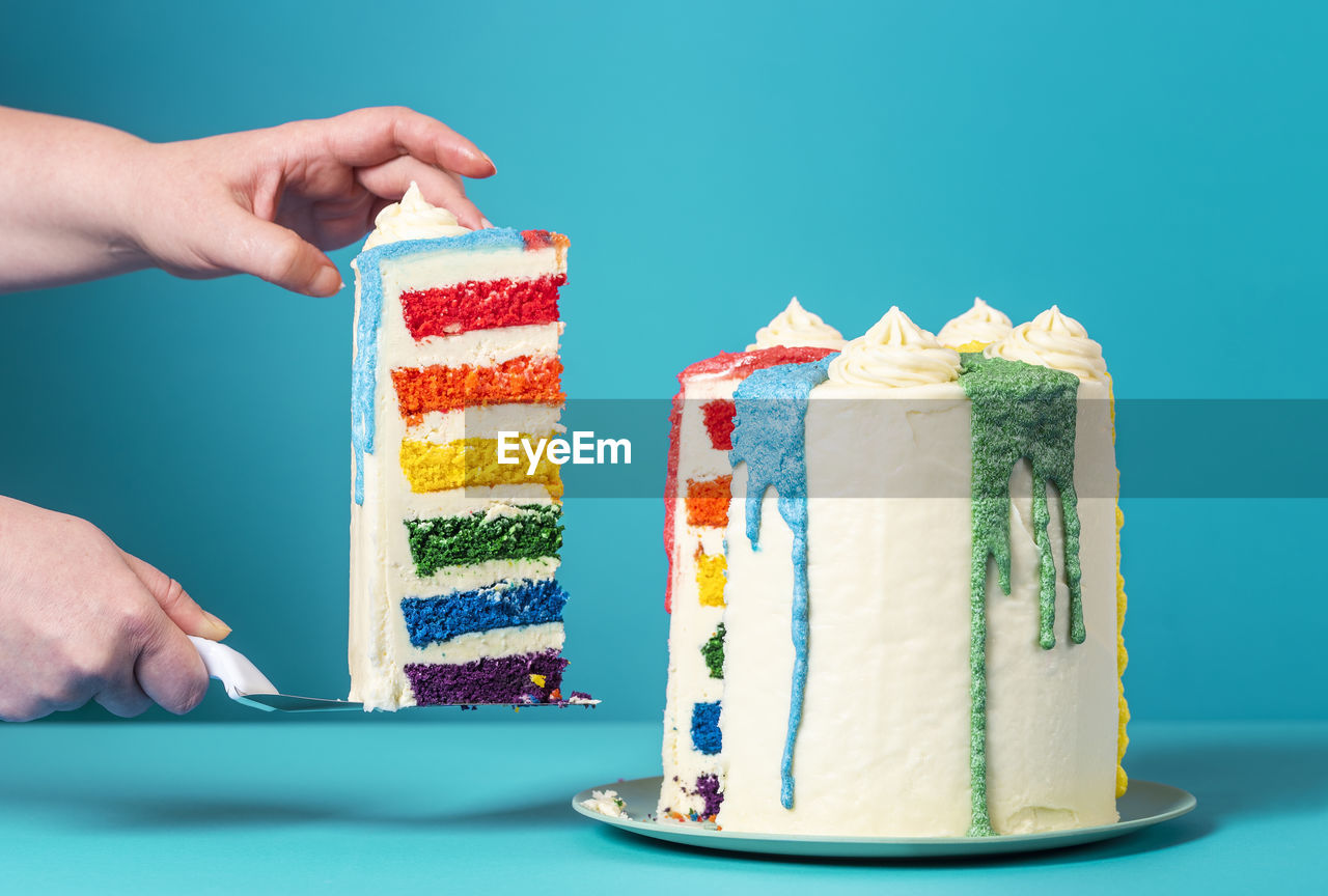 Woman's hands taking a slice of rainbow cake. homemade birthday cake against a blue background.