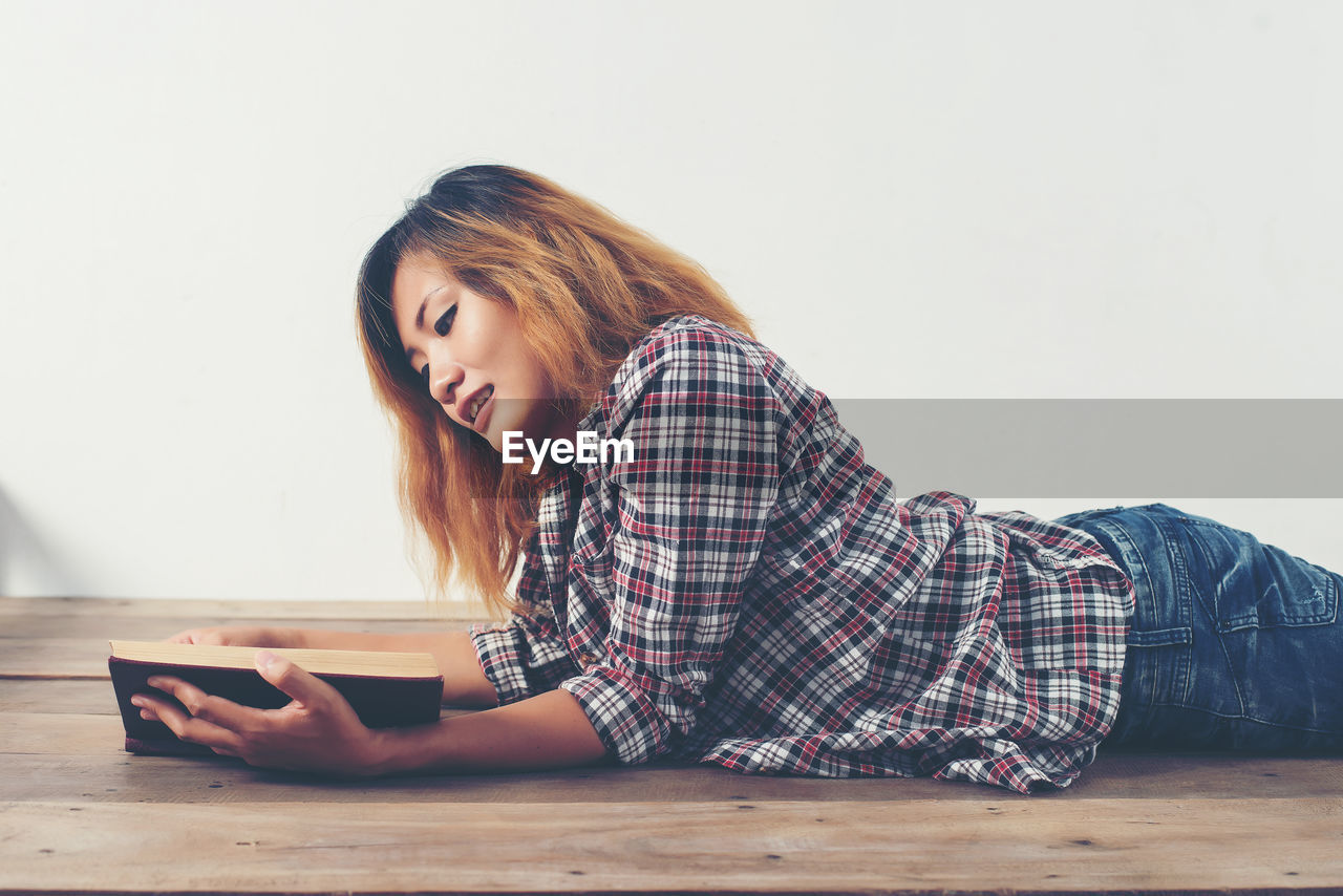 Smiling young woman reading book while lying on hardwood floor