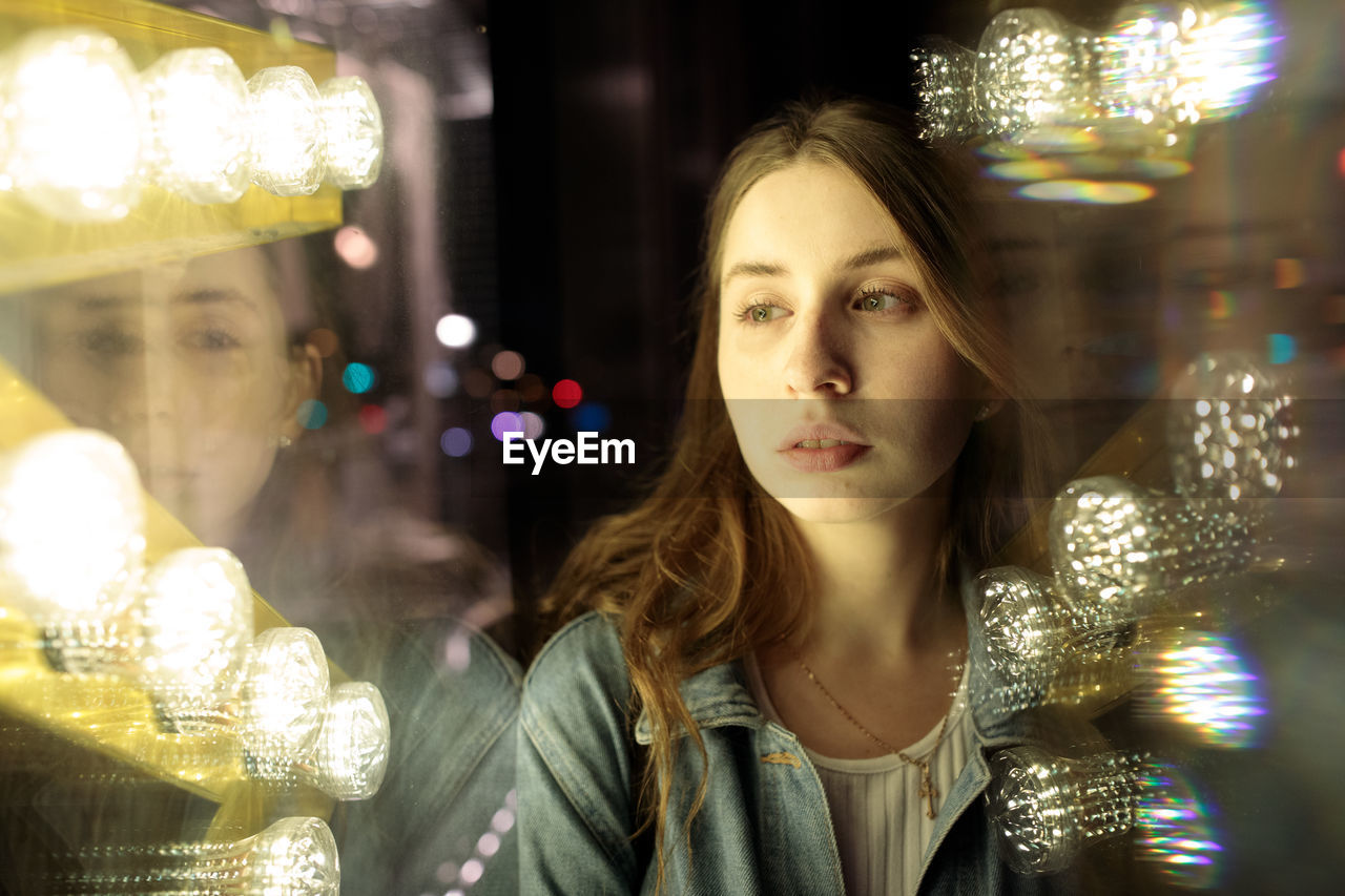 PORTRAIT OF YOUNG WOMAN LOOKING AT ILLUMINATED LIGHTS