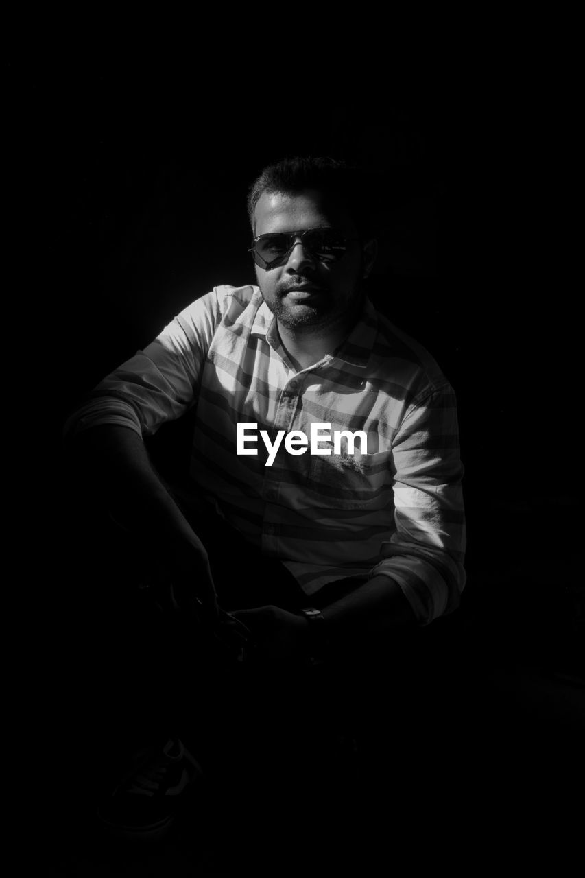 Portrait of young man wearing sunglasses while sitting in darkroom