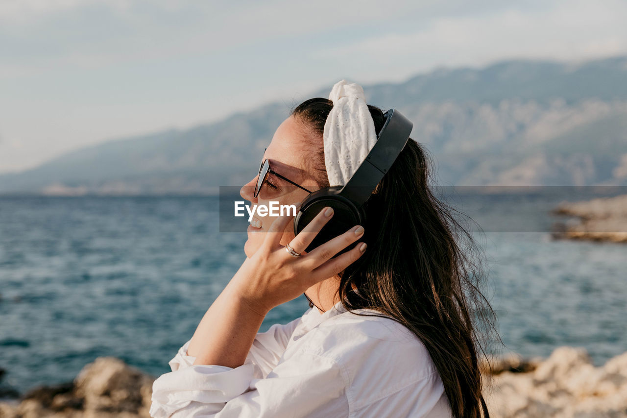 Side view of young woman in white shirt listening to music on headphones. summer, beach, lifestyle.